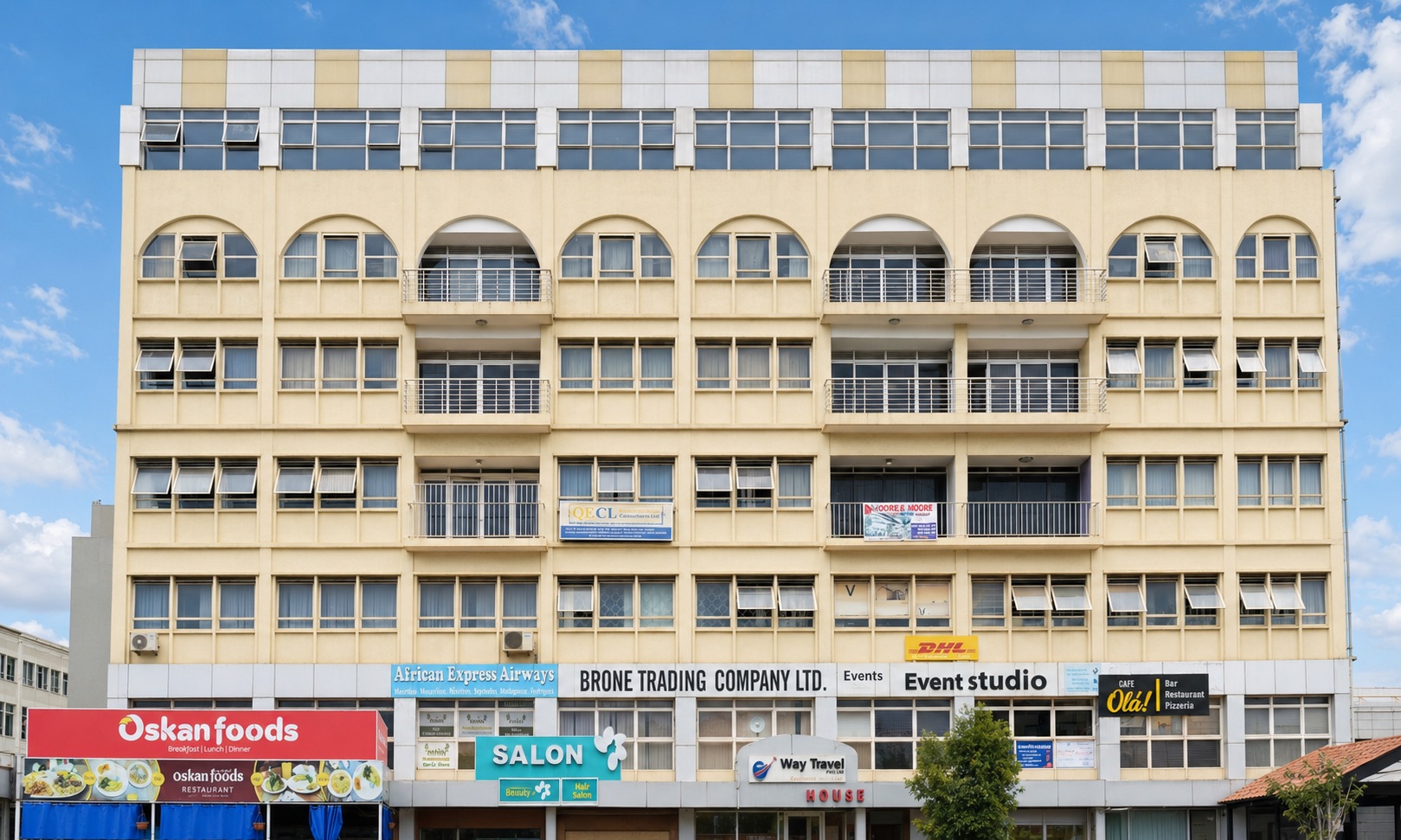 Shumuk House from across the street: a cream rendered building of eight levels, arched windows and balconies above, and a run of shopfronts along the pavement including Oskan foods, African Express Airways and Cafe Ola