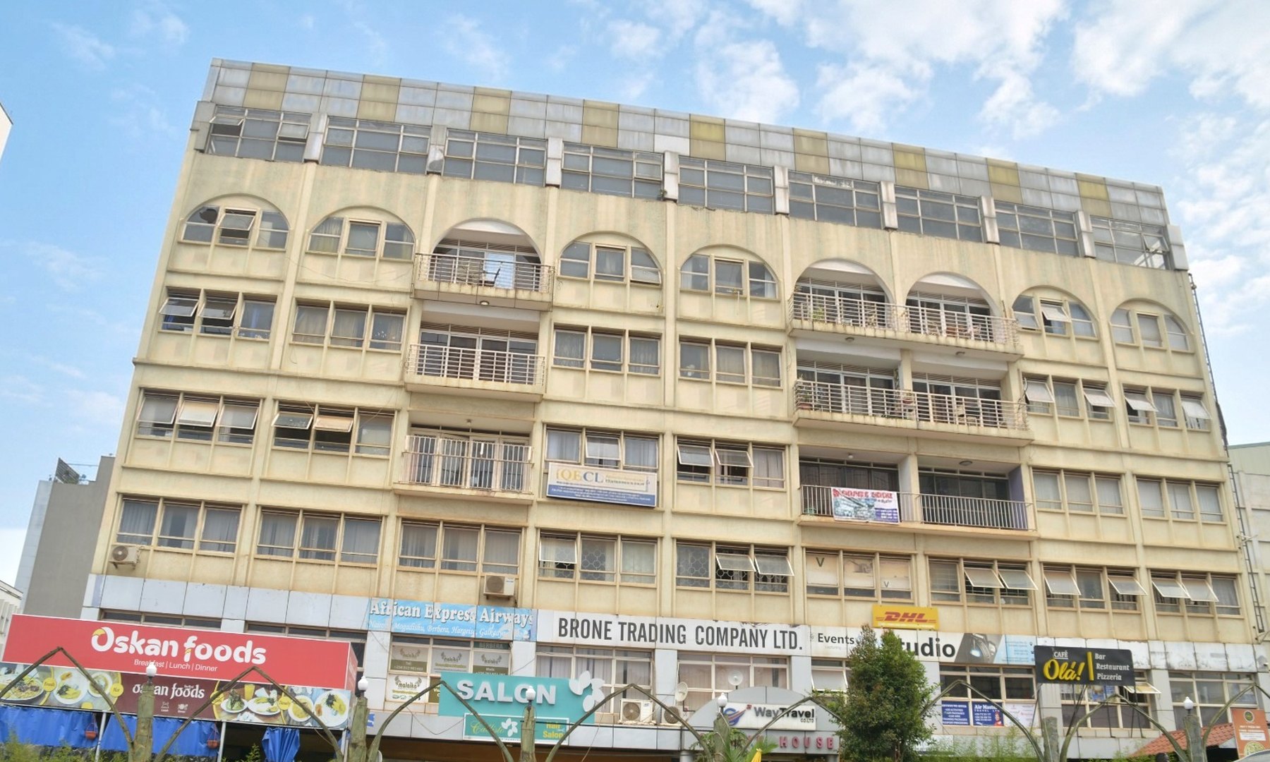 Shumuk House from across the street: a cream rendered building of eight levels, arched windows and balconies above, and a run of shopfronts along the pavement including Oskan foods, African Express Airways and Cafe Ola