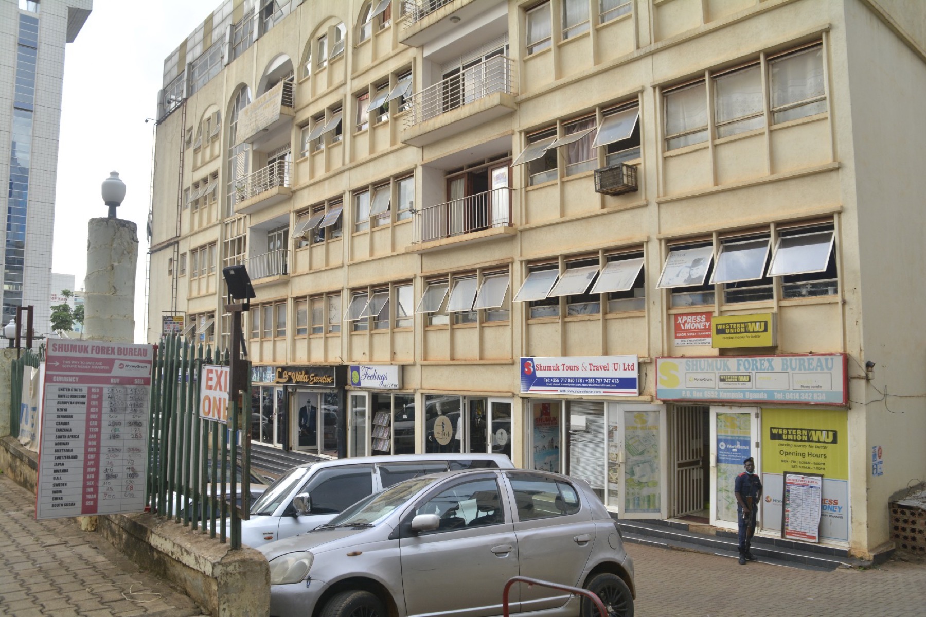 The pavement frontage of Shumuk House: the Shumuk Forex Bureau currency board, shopfronts either side, and a security guard at the entrance